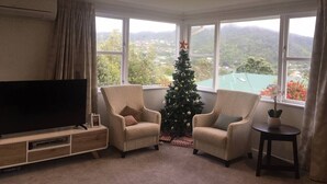 Un soggiorno con una TV a schermo piatto, due poltrone, un albero di Natale e vista su un paesaggio verde attraverso ampie finestre.
