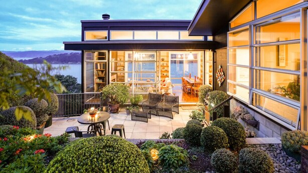 A modern house with a large glass door leading to a patio area with outdoor furniture and a view of the lake. - Roseneath Escape - Panoramic Harbour Views (Wellington)