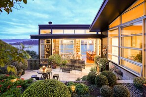 A modern house with a large glass door leading to a patio area with outdoor furniture and a view of the lake. - Roseneath Escape - Panoramic Harbour Views (Wellington)