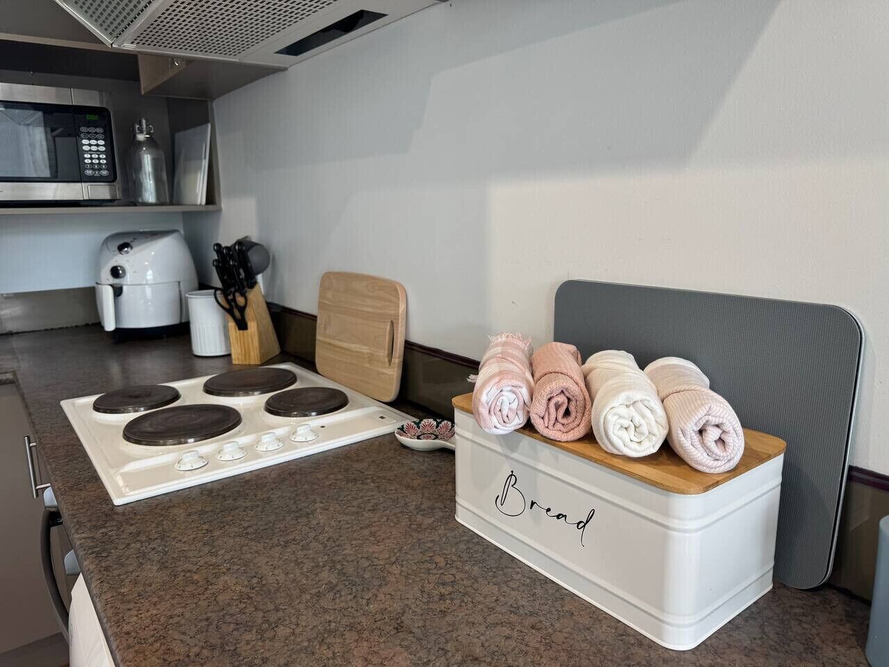 A kitchen with a stove, a coffee maker, a cutting board, and a bread box with towels.