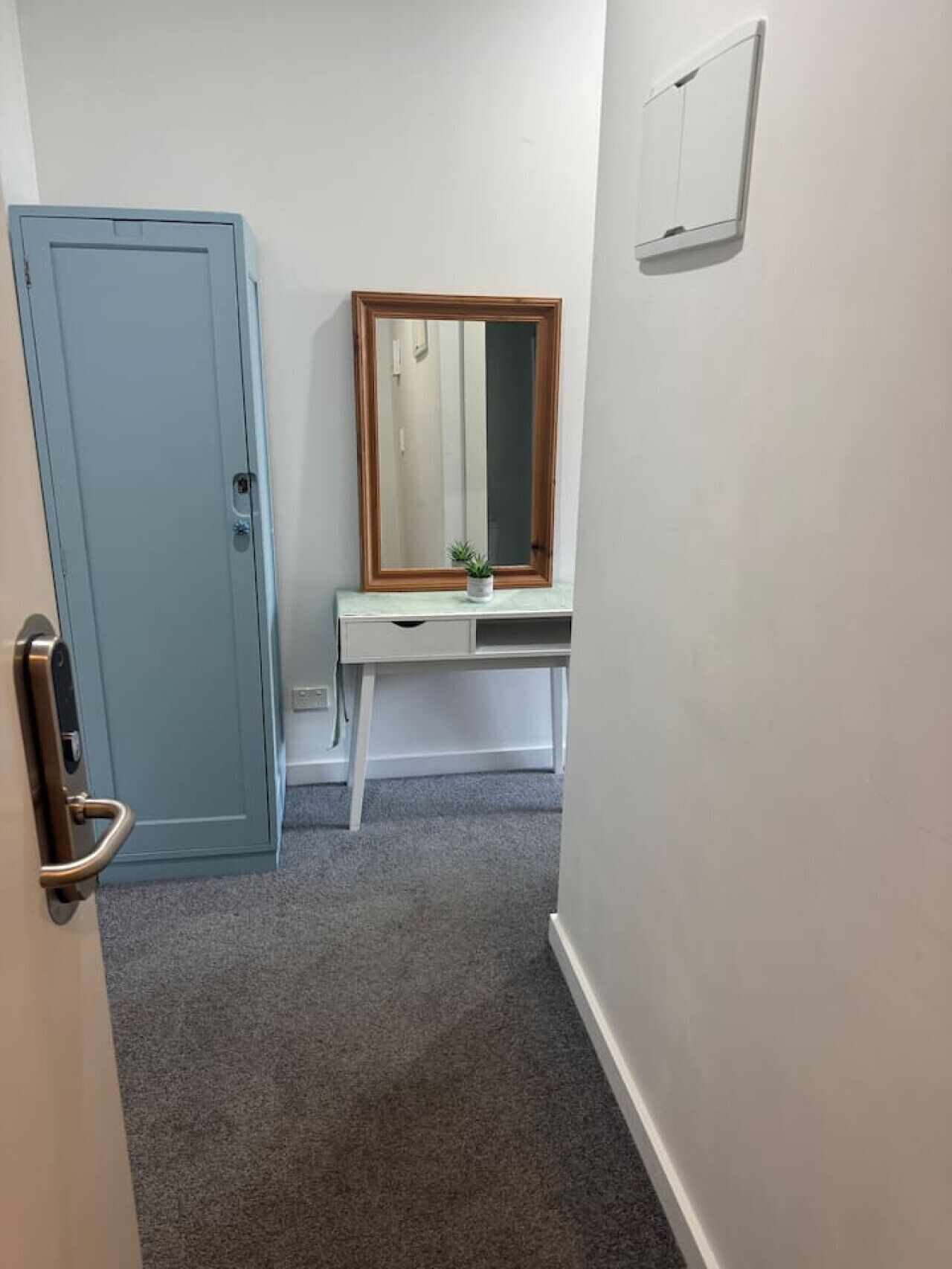 A narrow hallway with a blue door, a white desk with a mirror, and a small plant.