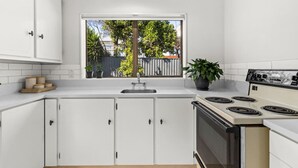 A modern kitchen with white cabinetry, a large window with a view of trees and a fence, and a stove with four burners. - Havelock North Heartbeat (Havelock North)