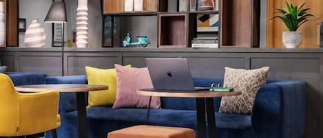 A modern living room with a blue sofa, yellow and pink cushions, a small table with a laptop, and a potted plant.