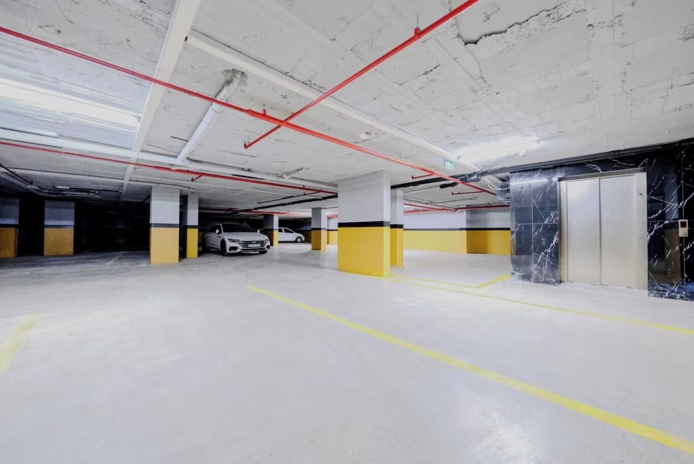 Un estacionamiento vacío con pilares amarillos y blancos, un coche blanco estacionado y un ascensor a la derecha.