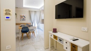 Un soggiorno moderno con una TV a schermo piatto, una zona pranzo con sedie e tavolo, e un cartello con i nomi delle stanze.