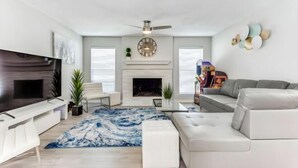 A modern living room with a flat-screen TV, a fireplace, a sofa, and a ceiling fan. - Reston NEW Bright, Airy Home w/ In Ground Pool & Grill (Richardson)