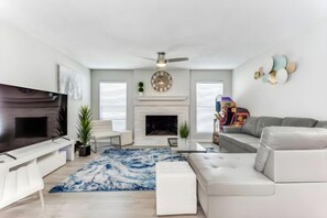 A modern living room with a flat-screen TV, a fireplace, a sofa, and a ceiling fan. - Reston NEW Bright, Airy Home w/ In Ground Pool & Grill (Richardson)