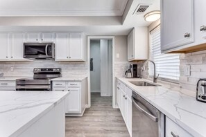 A modern kitchen with white cabinetry, a built-in microwave, a stove, and a large island with a sink. - Reston NEW Bright, Airy Home w/ In Ground Pool & Grill (Richardson)