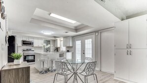 A modern kitchen with white cabinets, a dining area with a glass table, and a large wardrobe. - Reston NEW Bright, Airy Home w/ In Ground Pool & Grill (Richardson)