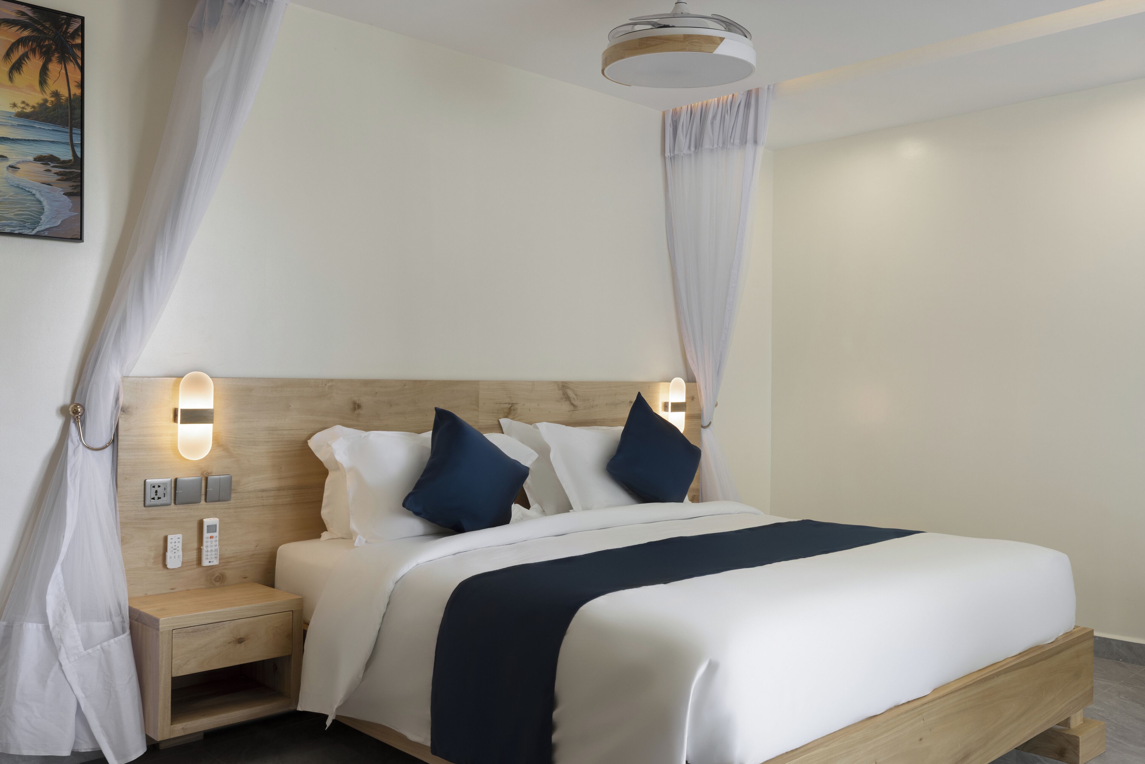 Una cama bien hecha con cabecera de madera, ropa de cama blanca y almohadas azul oscuro. Hay una mesita de noche con una lámpara y un enchufe.