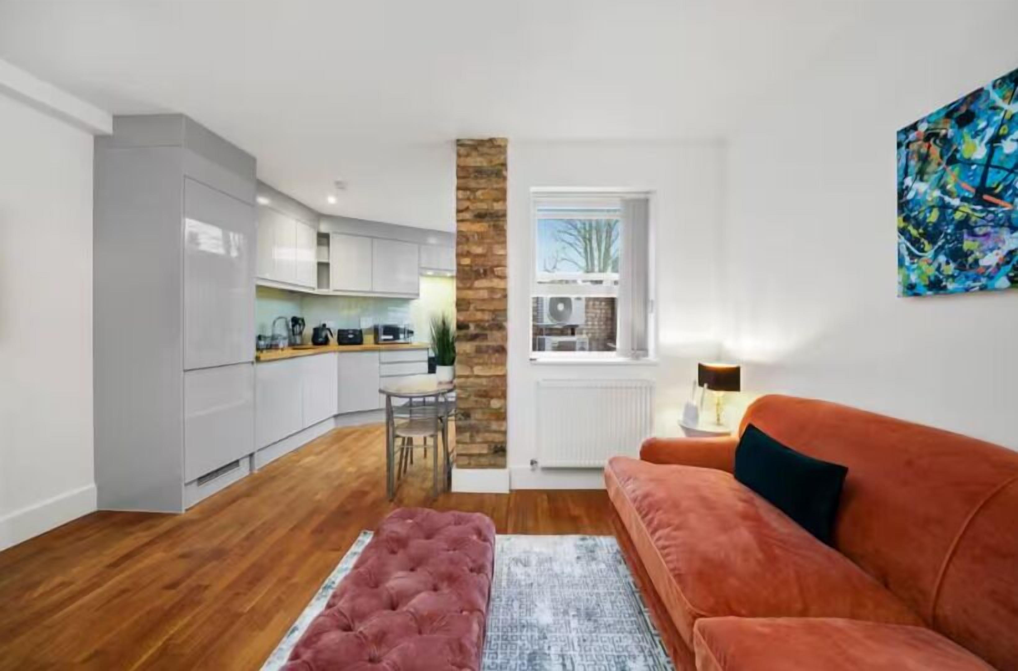 Un salon moderne avec un canapé, un pouf et un coin cuisine doté de meubles blancs et d’un mur d’accent en pierre.