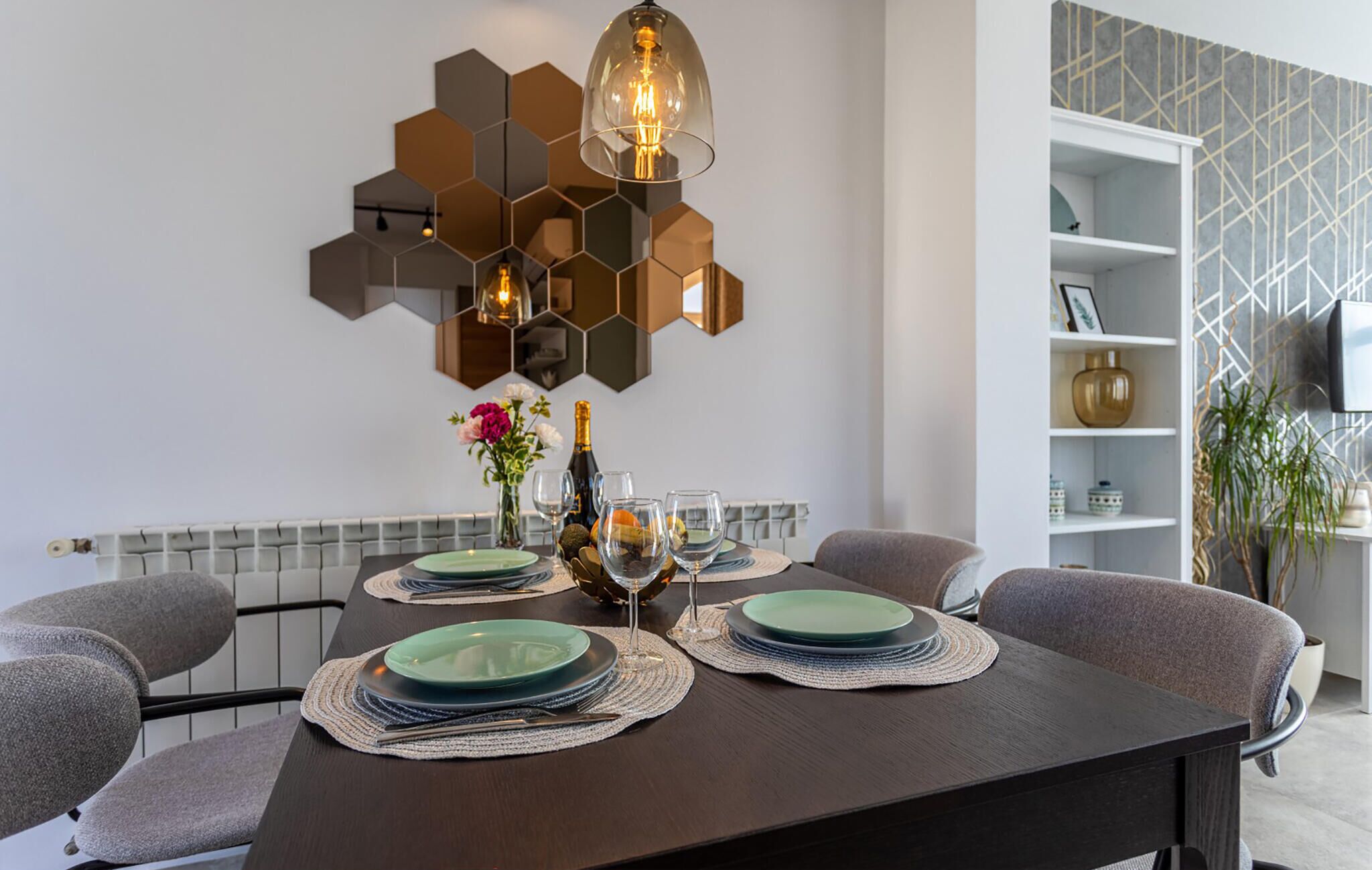 A dining area with a dark wooden table set for four, hexagonal wall decor, and a modern shelving unit with decorative items.