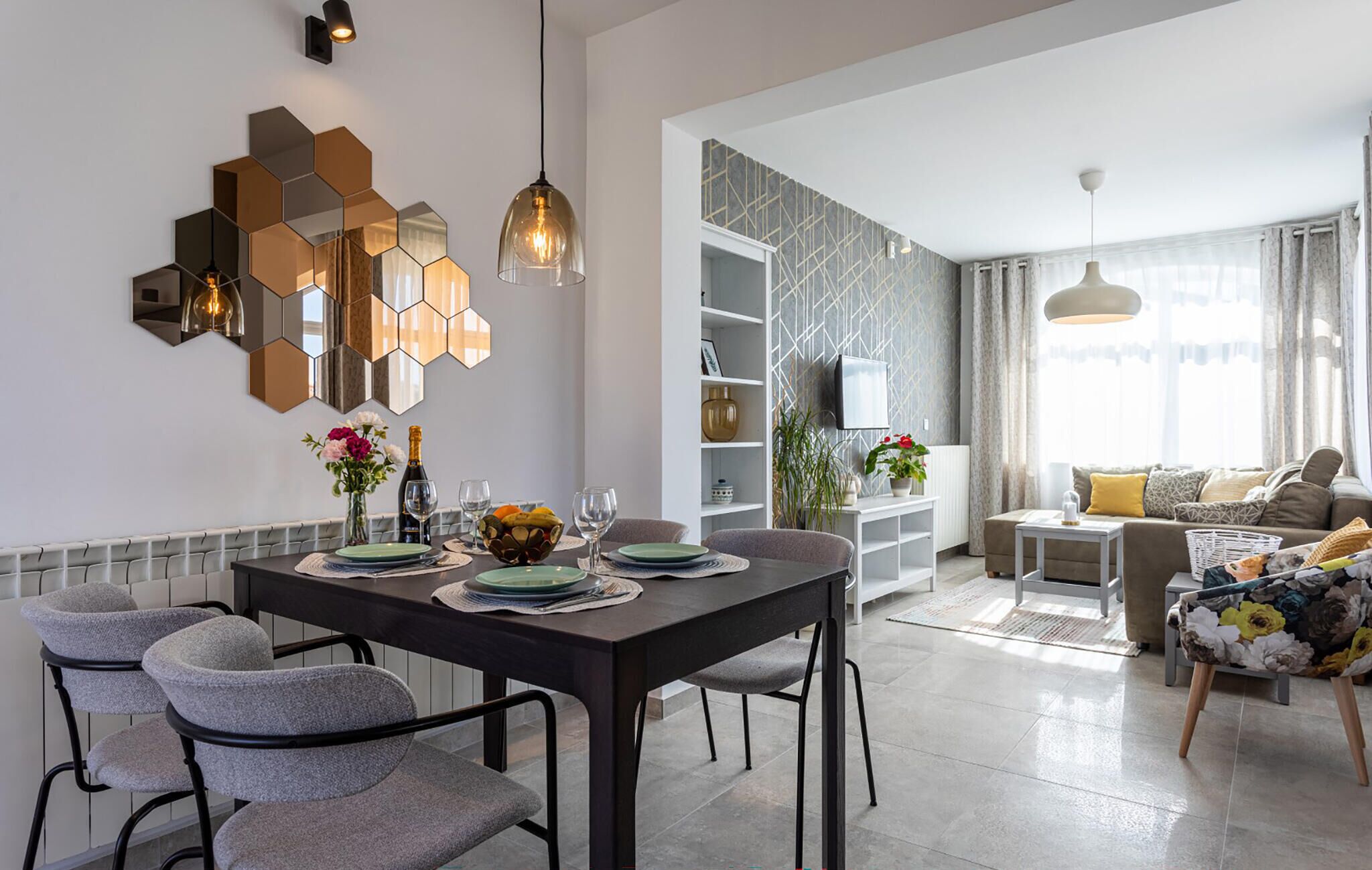 A modern living room with a dining area, featuring a hexagonal wall decoration, a flat-screen TV, and a sofa with a floral cushion.