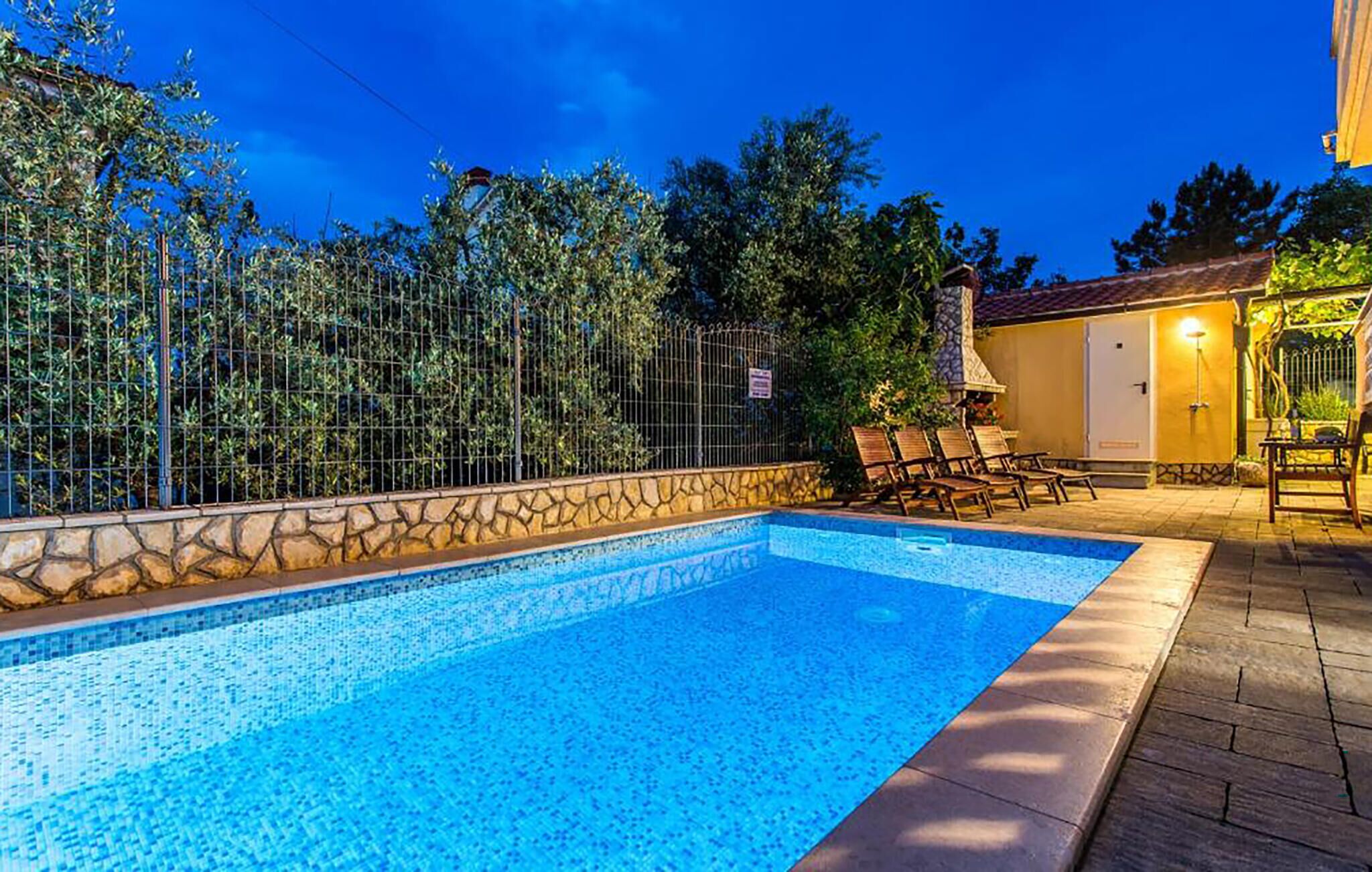 Una piscina con borde de piedra, rodeada por una valla y árboles. Se observan sillas de descanso y un edificio con una puerta y un aplique.