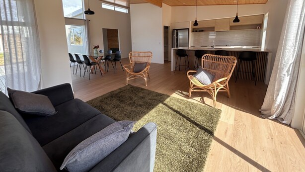 A modern living room with a sofa, two wicker chairs, a dining area with a table and chairs, a kitchen with bar stools, and a large window with curtains. - Mapua Haven – Contemporary New Home (Māpua)