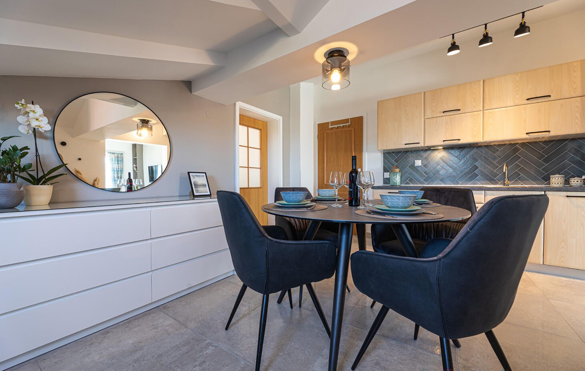 A modern kitchen with wooden cabinets, a herringbone backsplash, and a dining area with a round table and two chairs.
