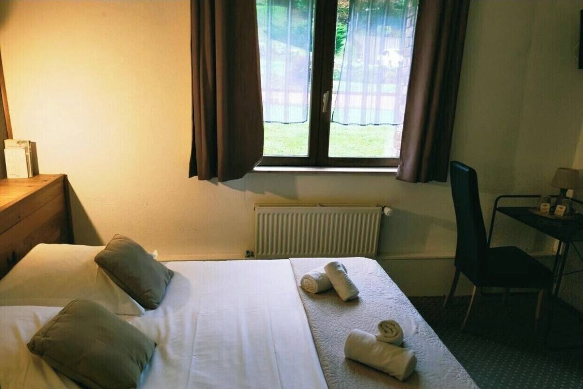 A hotel room with a bed, pillows, a desk, a chair, a radiator, and a window with curtains.