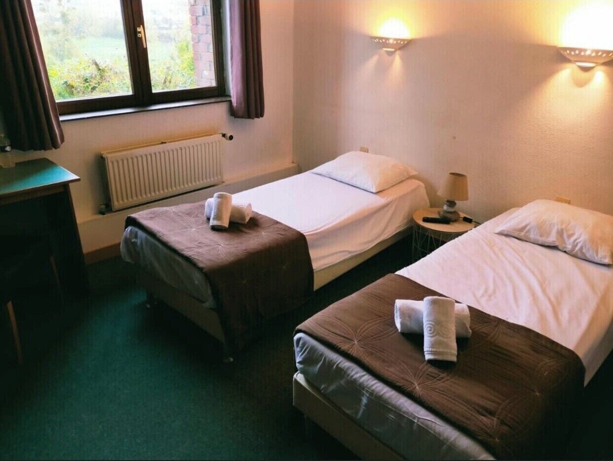 Een hotelkamer met twee bedden, een raam met gordijnen, een radiator en een nachtkastje met een lamp.