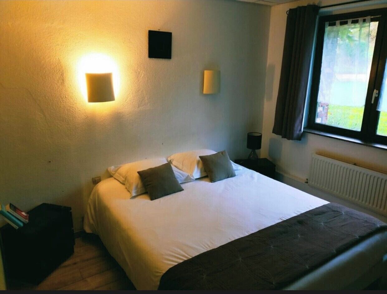 Un llit ben fet amb almoades, una taula de nit amb una làmpada, i una finestra amb cortines.