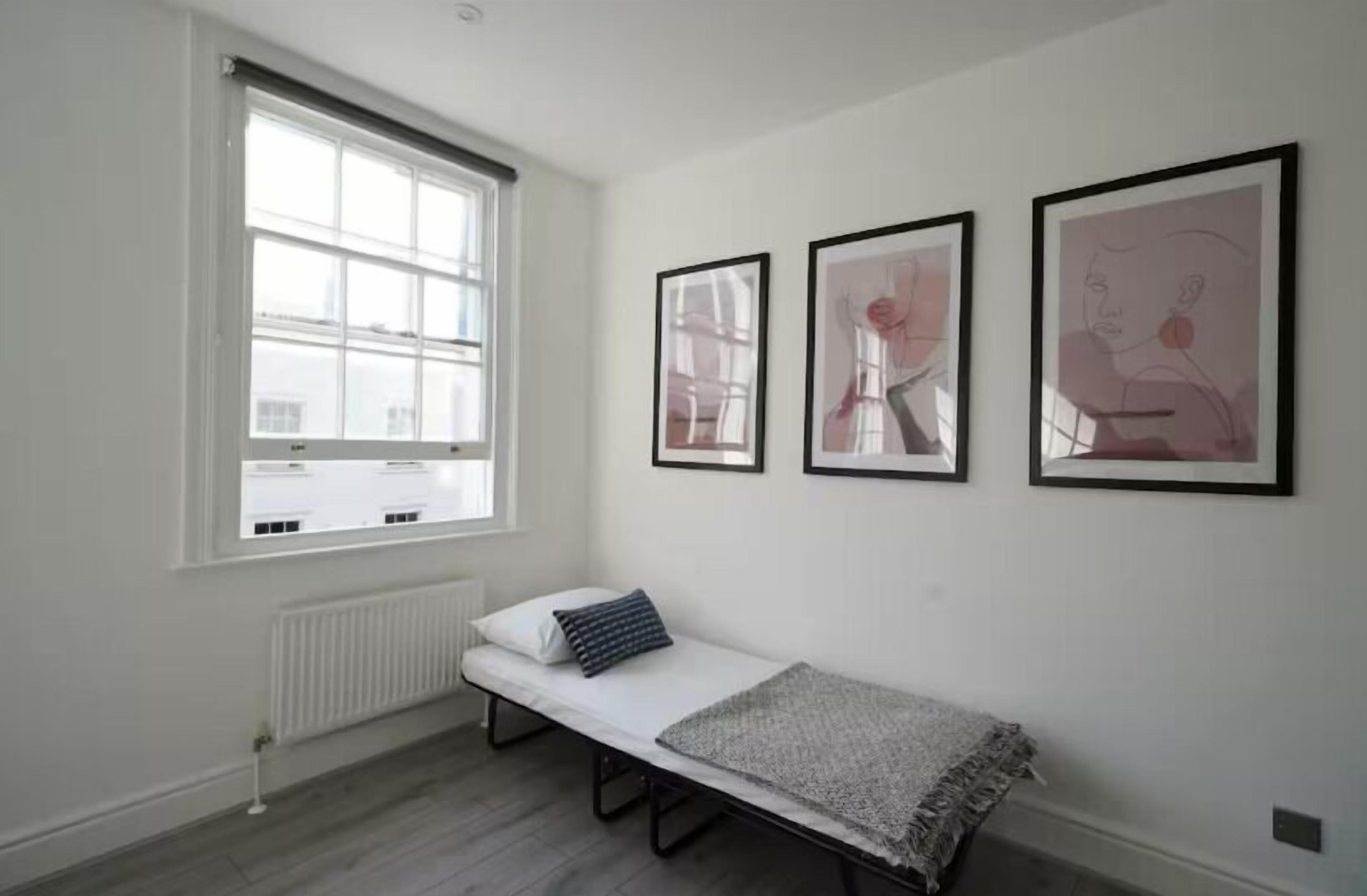 A small, single-bed room with a window, radiator, and three framed artworks on the wall.