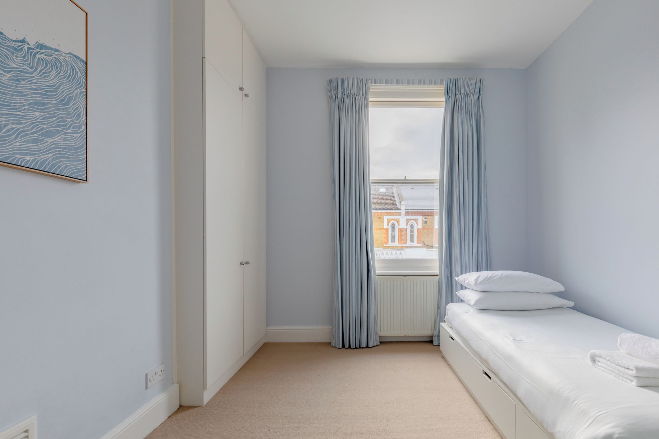 A bedroom with a single bed, a window with curtains, a radiator, and a wall-mounted light switch.
