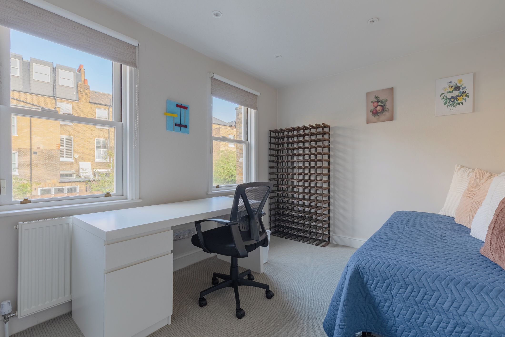 A bedroom with a white desk, a black office chair, a blue bed, and a window with a view of buildings.