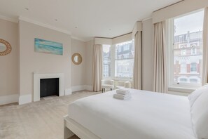 A hotel room with a large bed, a fireplace, a view of buildings through the window, and a painting on the wall. - Impeccably Designed 5BD Family Home in West London (Greater London)