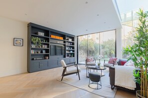 A modern living room with a large bookshelf, a sofa set, and a dining area. - Impeccably Designed 5BD Family Home in West London (Greater London)