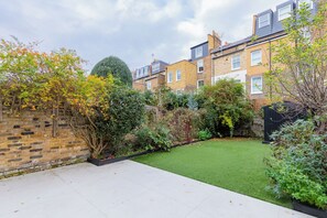 A residential garden with a lawn, brick wall, and mature trees. - Impeccably Designed 5BD Family Home in West London (Greater London)