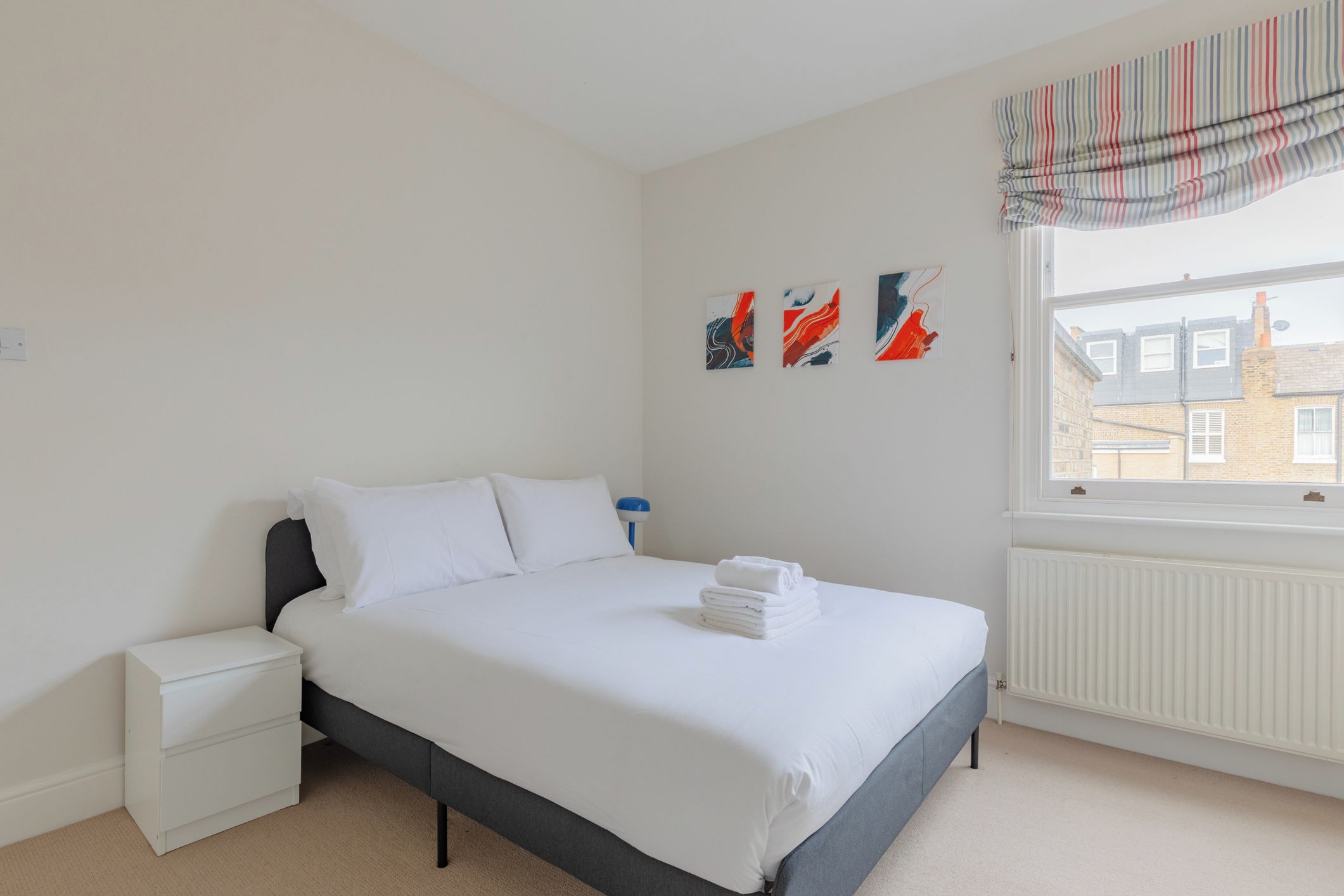 Une chambre à coucher avec un lit, une table de chevet, un radiateur et une fenêtre donnant sur des immeubles.