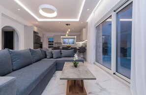 A modern living room with a grey sectional sofa, a wooden coffee table with a flower, and a large sliding glass door to the right. - Villa Orizzonte- First Floor Apartment (Zakynthos)