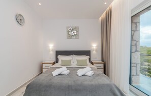 A modern bedroom with a large bed, two bedside tables, a wall clock, and a window with curtains. - Villa Orizzonte- First Floor Apartment (Zakynthos)