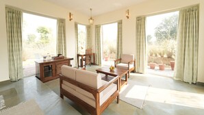 A spacious living area with a sofa, coffee table, and chairs. There is a wooden cabinet and a dining table with chairs. Large windows with curtains allow natural light and views of the outdoors. - The Rann Baagh Ranthambore (Sawai Madhopur)