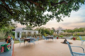 Un patio al aire libre con mesas y sillas, rodeado de follaje y un pequeño hogar para fuego.