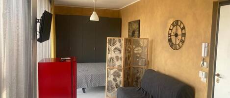 Una habitación de hotel con una cama, un armario rojo, un sofá gris, una mesa blanca y una planta decorativa.