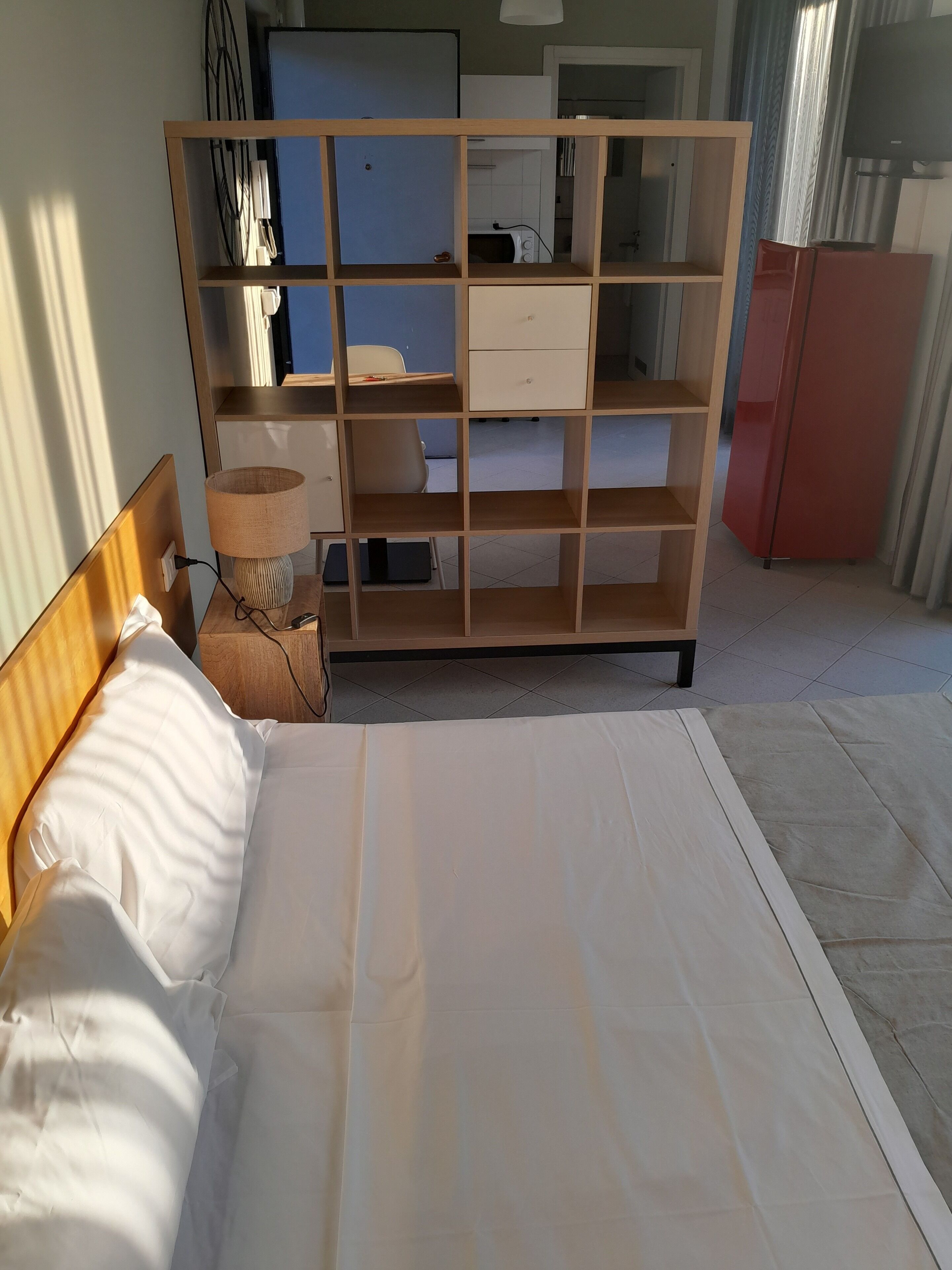 A hotel room with a bed, a wooden wardrobe, a desk with a lamp, and a red refrigerator.