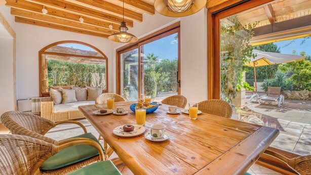 Un comedor con mesa de madera dispuesta para el desayuno, sillas de mimbre y grandes ventanales que ofrecen vista al jardín.