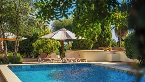 Piscina con sillones de descanso y sombrillas, rodeada de vegetación.