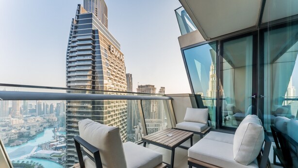 A balcony with two white cushioned chairs and a small wooden table, overlooking a cityscape with tall buildings and a river. - High Floor with Direct Burj Khalifa and Fountain Views (Dubai)