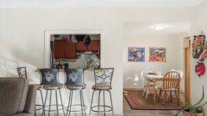 A modern living room with a sofa, two bar stools, a dining table with chairs, a kitchen area with cabinets and appliances, and two framed artworks on the wall.