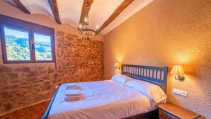 A bedroom with a stone wall, a wooden bed frame, a nightstand with a lamp, and a window with a view of the outdoors.