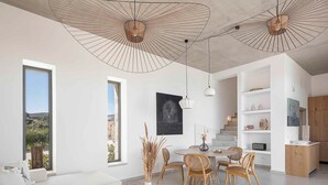 A modern living room with a white sofa, a round coffee table, and a dining area with wicker chairs. There are two pendant lights and a large window offering a view of the outdoors. - Himmel Villa I - Private pool, seaview&playground (Chania)