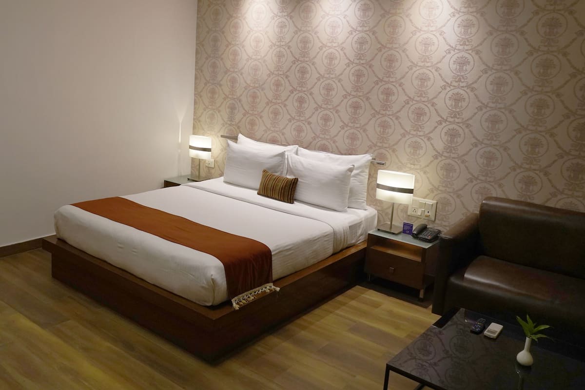 A hotel room with a bed, a brown leather sofa, a small table with a vase, and a patterned wallpaper.
