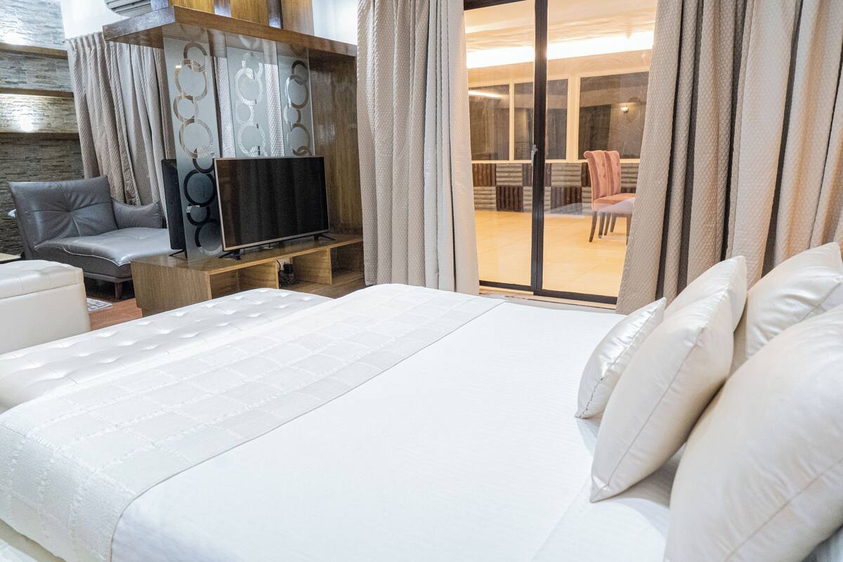 A hotel room with a large bed, a TV on a stand, a sofa, and a sliding glass door leading to a balcony.
