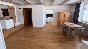 A spacious room with a wooden floor, a dining table with chairs, a wooden wardrobe, and a view of a kitchen through an open door.