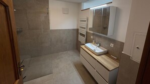 A modern bathroom with a walk-in shower, a wooden vanity, a large mirror, and a recessed ceiling light.