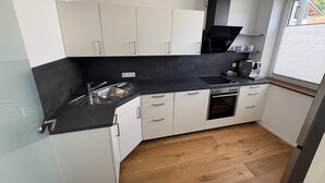 A compact kitchen with white cabinets, a black countertop, a built-in oven, and a window with blinds.