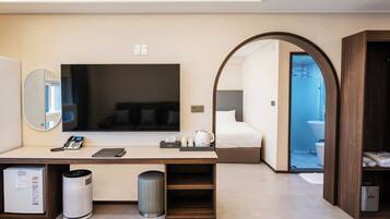 Una camera d'albergo moderna con una grande televisione a schermo piatto, una scrivania con telefono e laptop, uno specchio, un letto con biancheria bianca e vista sul bagno.
