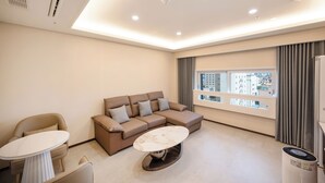 A modern living room with a sectional sofa, a round coffee table, and a view of the city through large windows. - VAVANT TOURIST HOTEL (Mokpo)