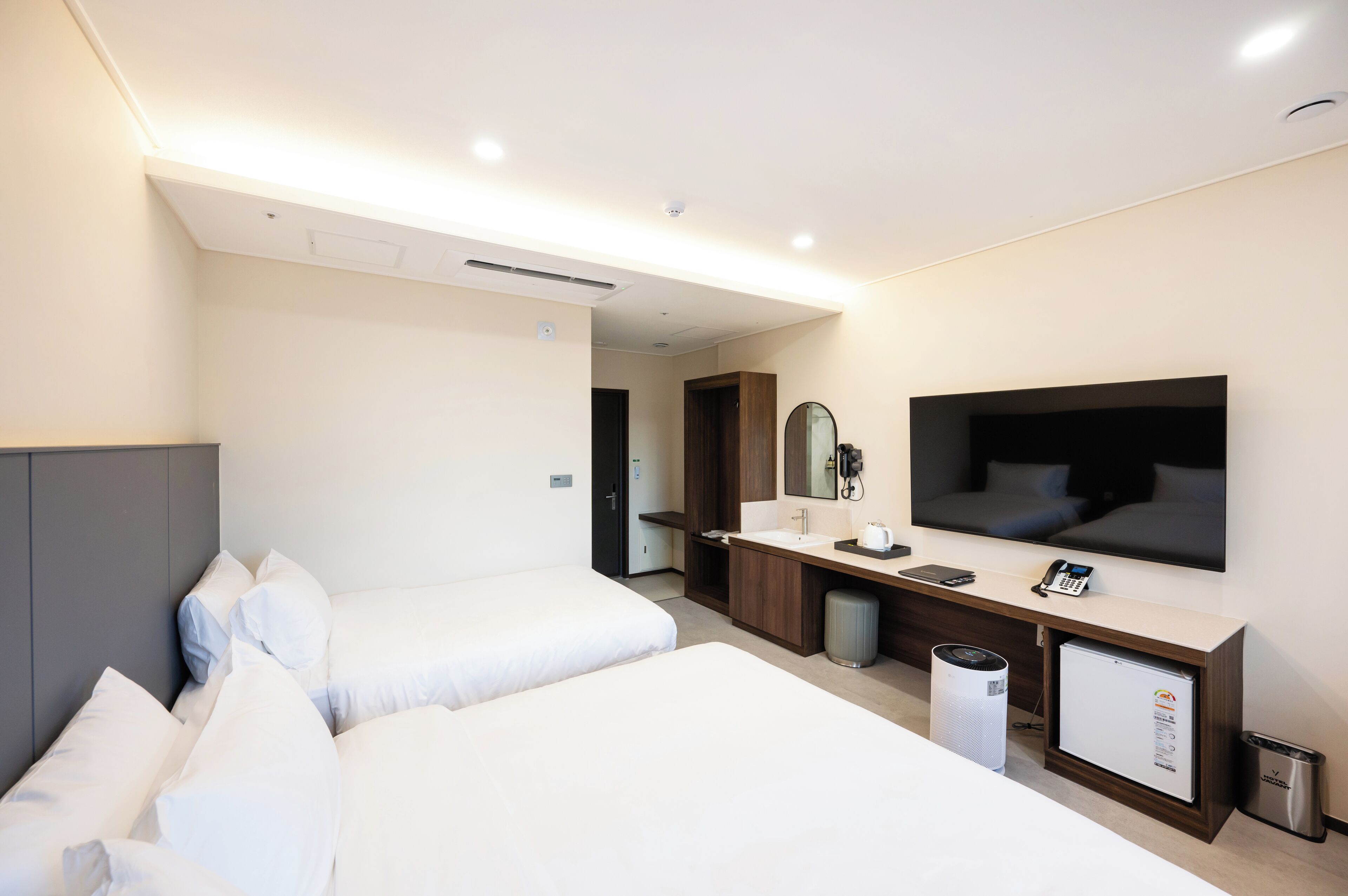 Quarto de hotel com duas camas, um espelho grande, uma TV, uma pequena geladeira e uma mesa com telefone e luminária.